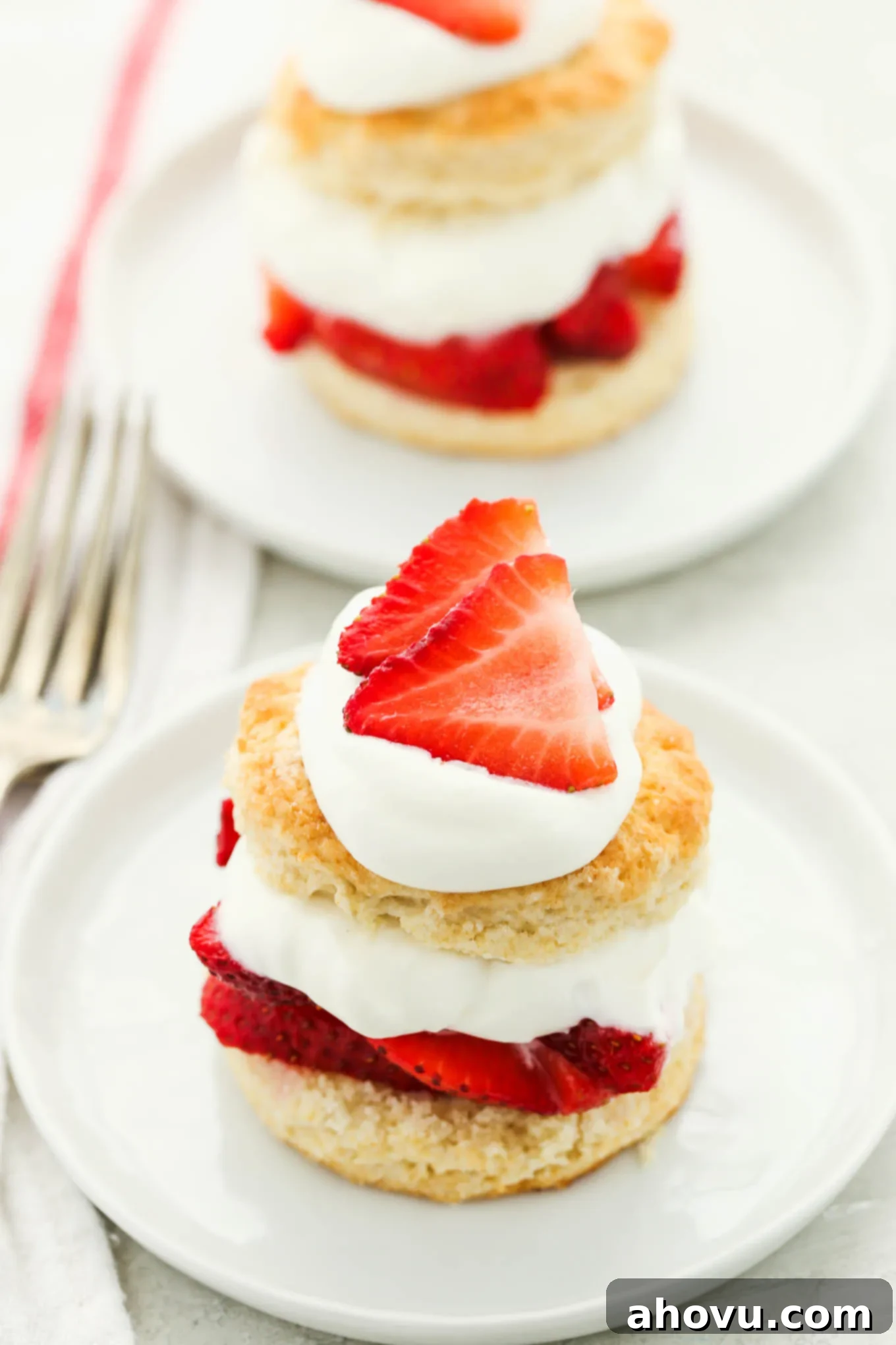 Two prepared strawberry shortcakes topped with extra whipped cream and sliced strawberries on white plates, showcasing the layers of biscuit, berries, and cream.