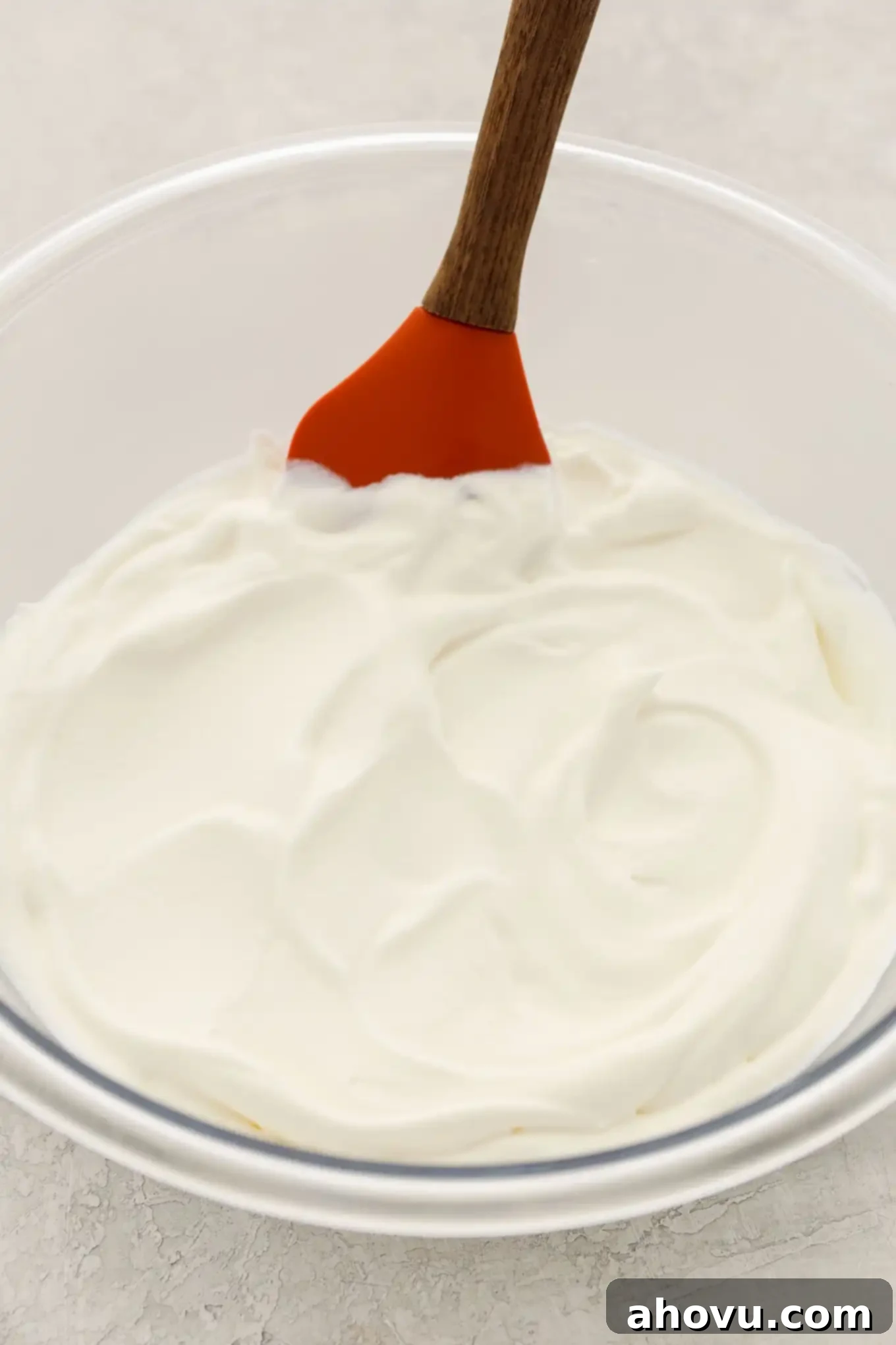 A clear glass bowl brimming with freshly made, fluffy homemade whipped cream, with a rubber spatula resting in it, ready to top a delicious dessert.