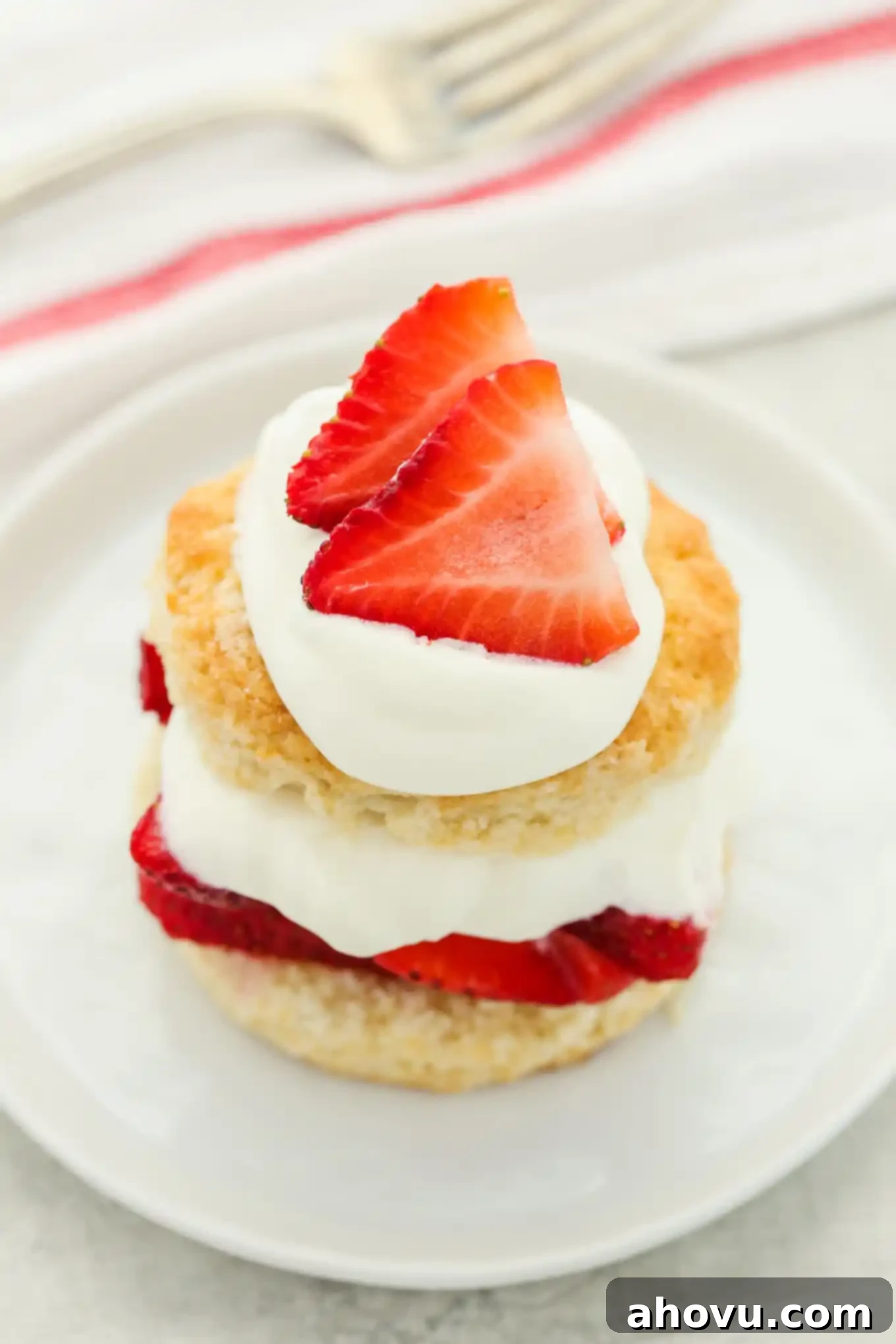 A perfectly assembled strawberry shortcake, generously topped with extra whipped cream and beautifully sliced strawberries, resting on a pristine white plate.