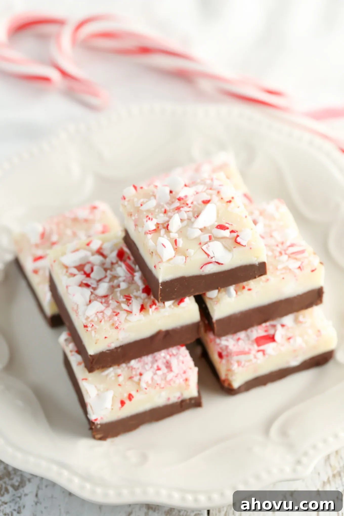 A close-up stack of colorful Christmas peppermint fudge on a white plate with two whole candy canes in the soft-focus background.
