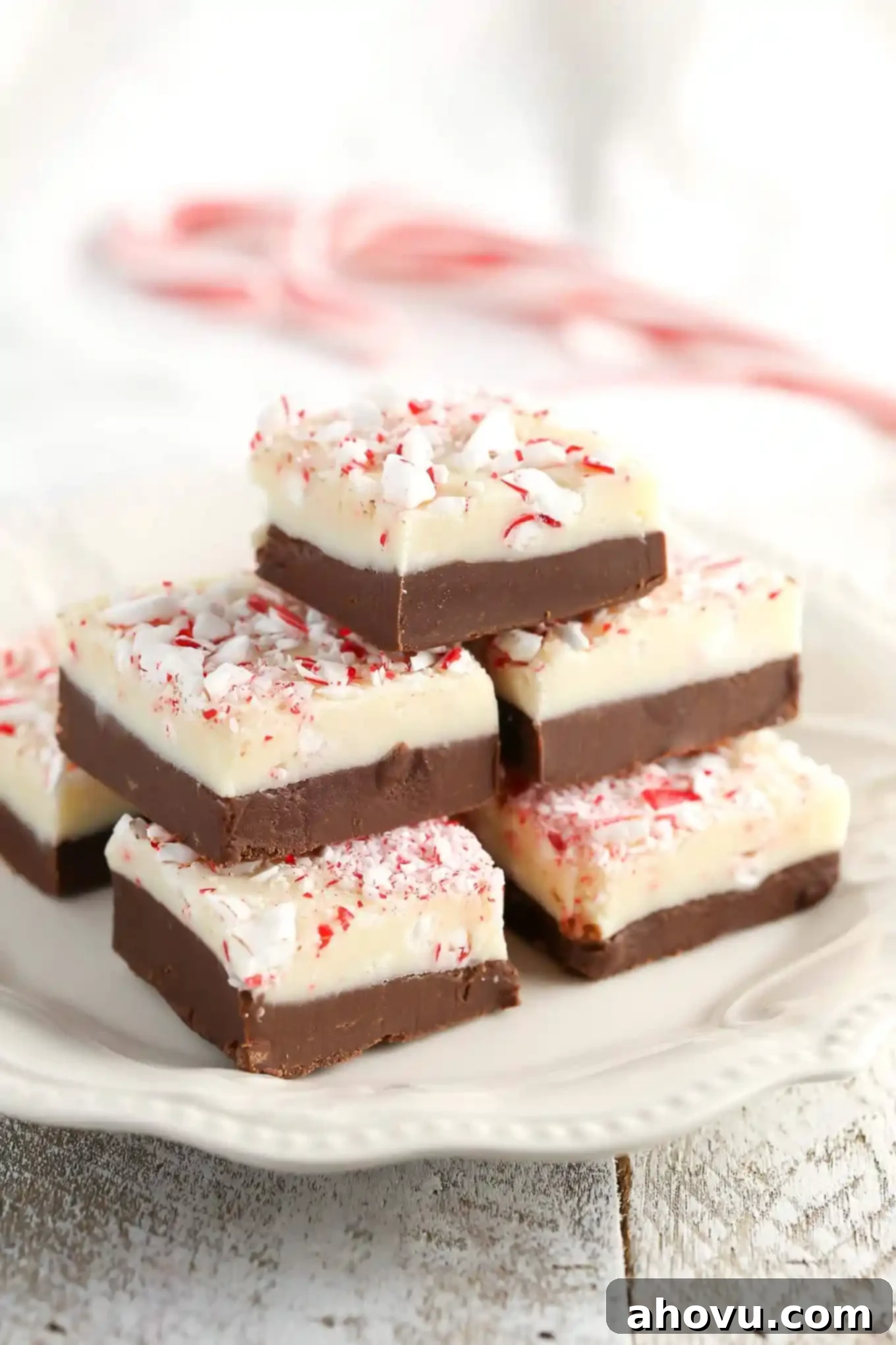 A stack of peppermint fudge squares on a white plate, showcasing the two distinct layers and candy cane topping.