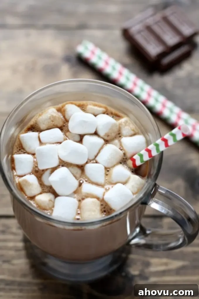 Mug of homemade hot chocolate topped with mini marshmallows and a straw. Chocolate squares rest in the background. 