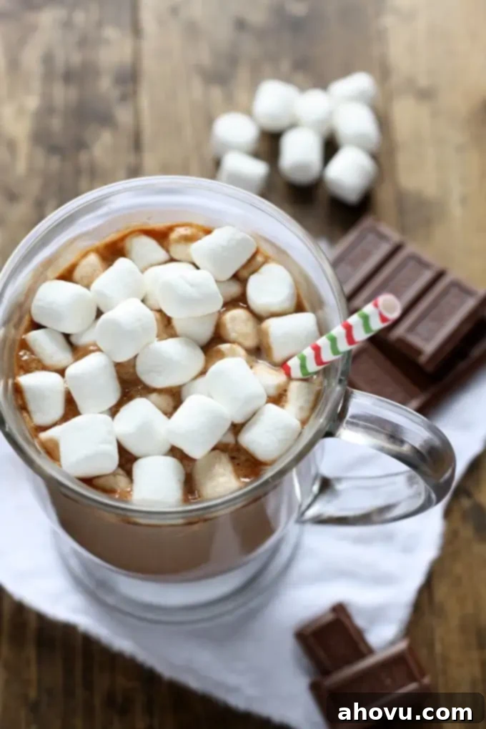 Mug of homemade hot chocolate topped with mini marshmallows and a straw. Chocolate squares and marshmallows rest in the background. 