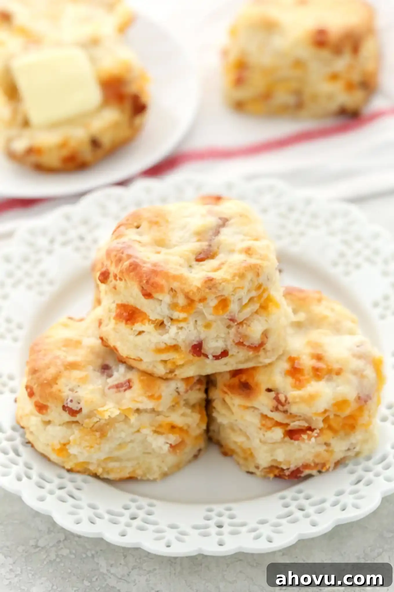 A stack of three golden-brown bacon cheddar biscuits on a white plate. Additional biscuits rest in the background, showing their flaky layers.