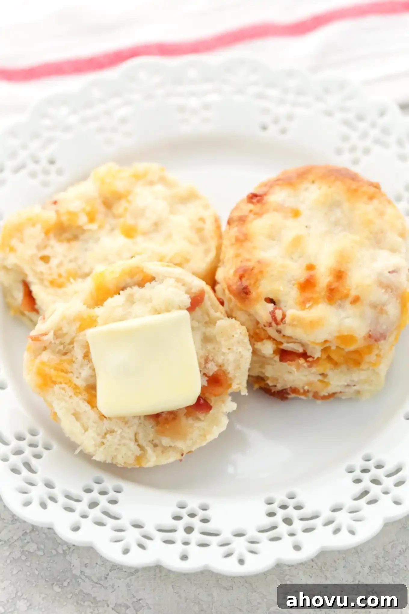 Two warm bacon cheddar biscuits on a white plate. One biscuit is sliced in half, revealing its flaky interior and a pat of melting butter.