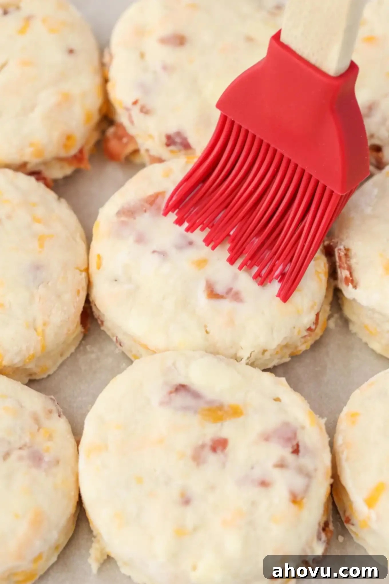 Three rows of unbaked cheddar cheese biscuits arranged on a baking sheet, being brushed with buttermilk for a golden finish.