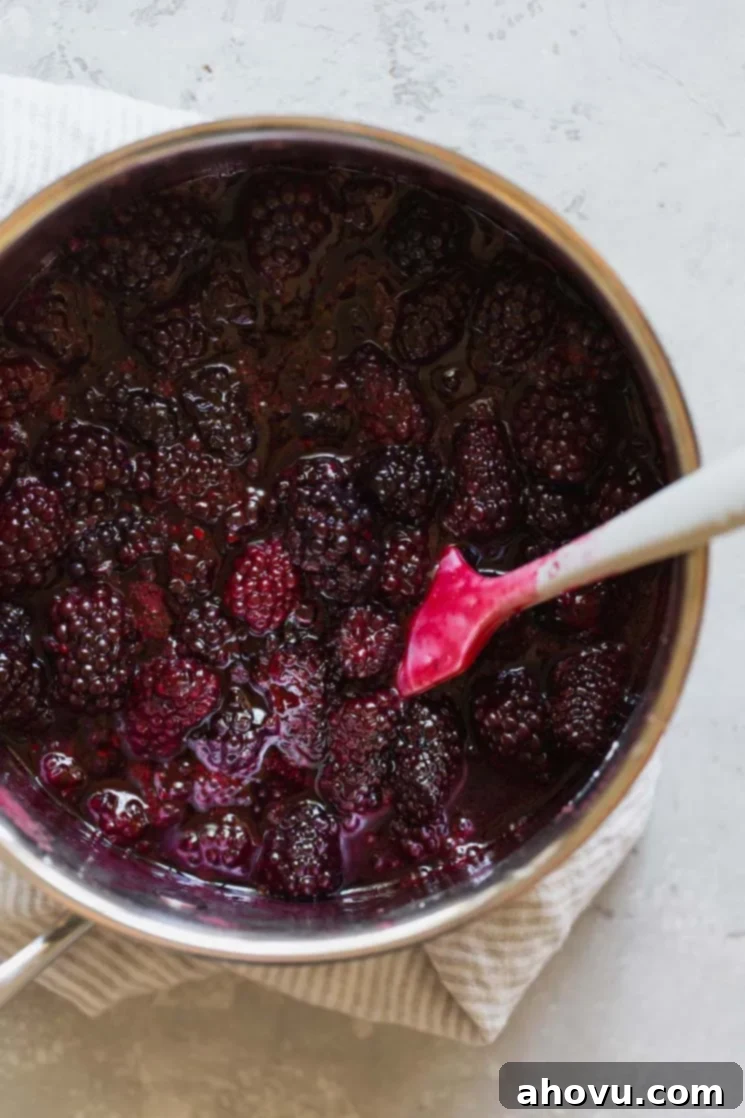 A close-up shot of a saucepan filled with vibrant blackberry filling, actively simmering and being stirred with a white spatula to achieve the perfect thickened consistency.
