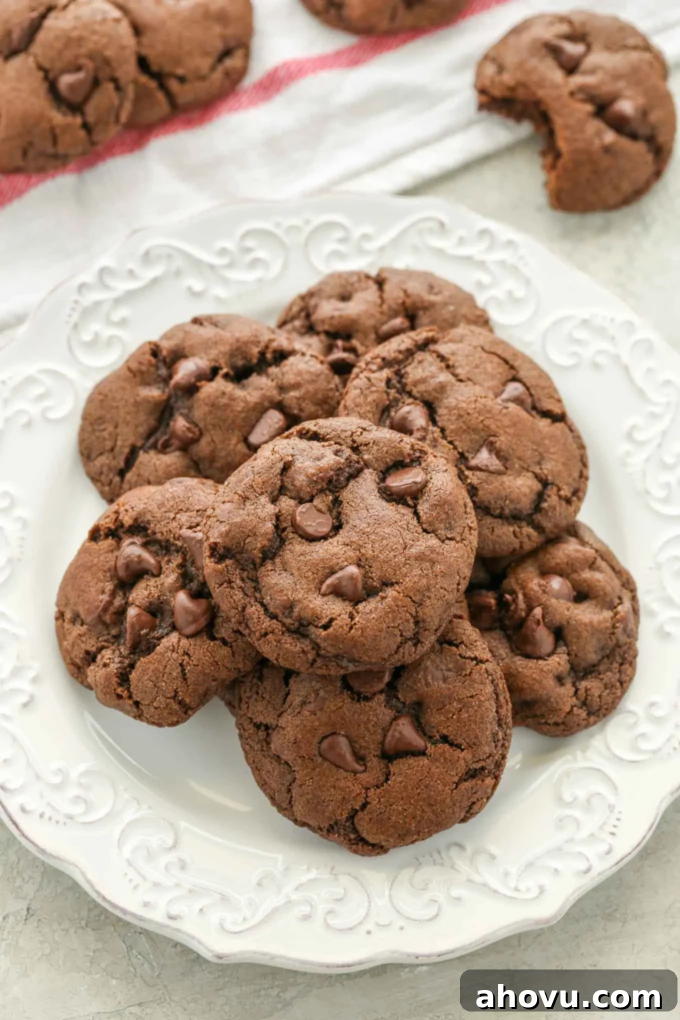 A stack of warm, soft double chocolate chip cookies on a white plate, with more cookies casually arranged around it, inviting to be eaten.