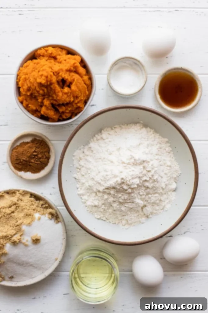 Various ingredients for pumpkin cake arranged on a rustic white wooden surface, including pumpkin puree, spices, flour, sugar, and eggs.