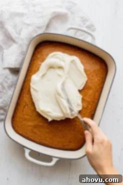 A creamy, white cream cheese frosting being generously spread over the top of a freshly baked pumpkin cake in a pan.