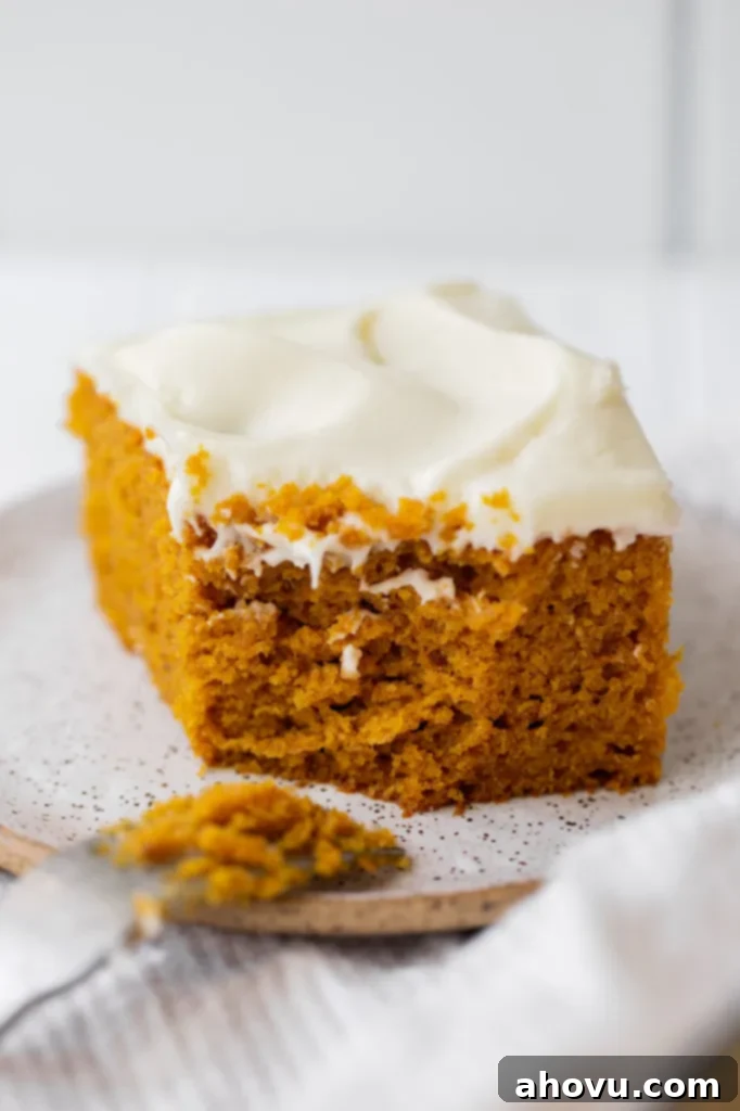 A single slice of moist pumpkin cake with cream cheese frosting on a speckled white plate, with a bite taken out, showing the cake's texture.