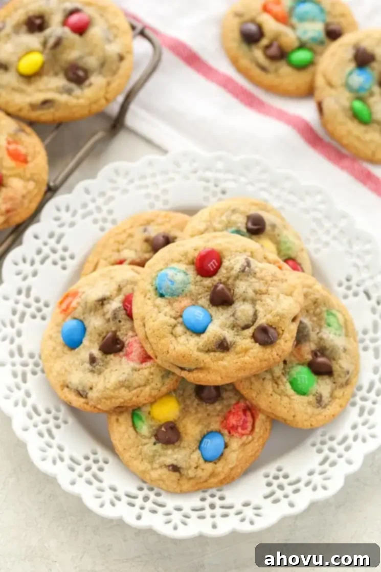 A white plate piled high with chocolate chip M&M's cookies. Additional cookies rest in the background. 