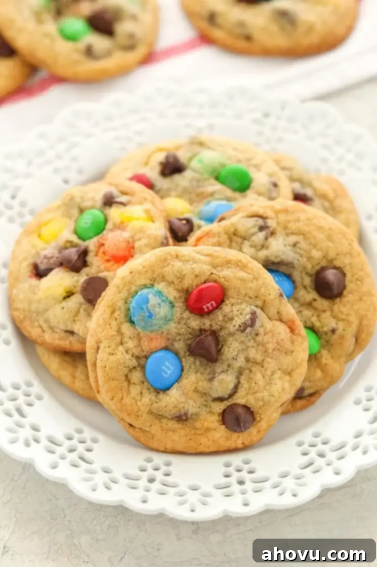 A pile of M&M's cookies on a white plate. Additional cookies rest in the background. 