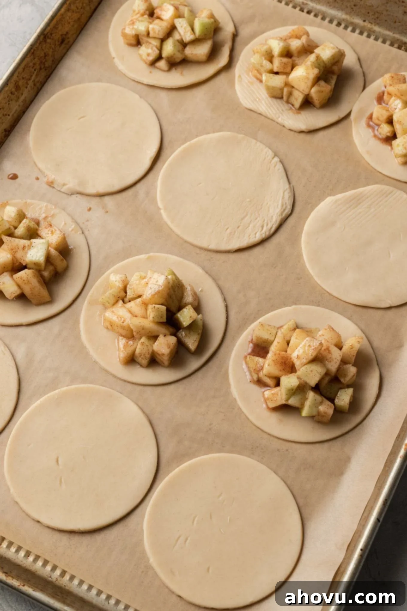 Flaky Apple Turnovers 5 A process shot demonstrating the assembly of apple hand pies, with circles of pie dough neatly arranged on a baking sheet, each topped with a mound of apple pie filling.