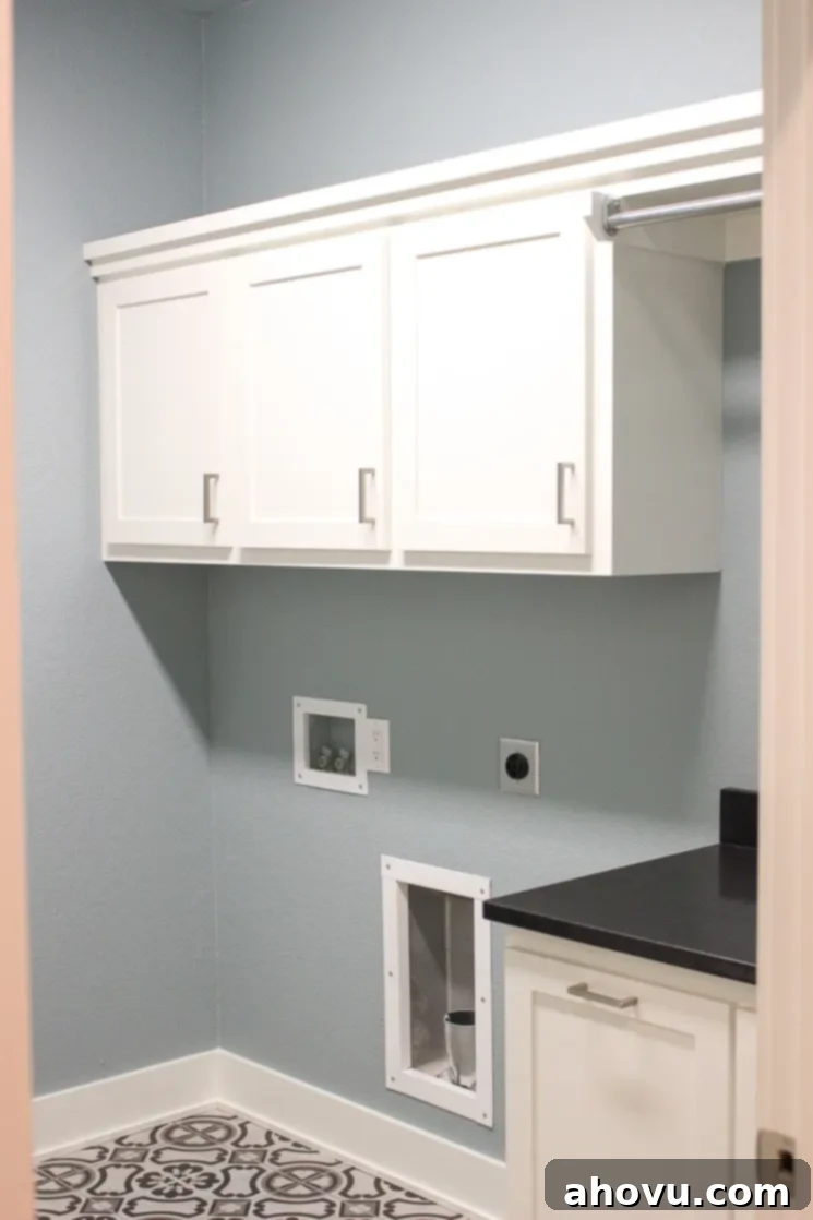 A functional laundry room featuring serene blue walls, bright white cabinets, and a gray porcelain tile floor designed to mimic concrete.