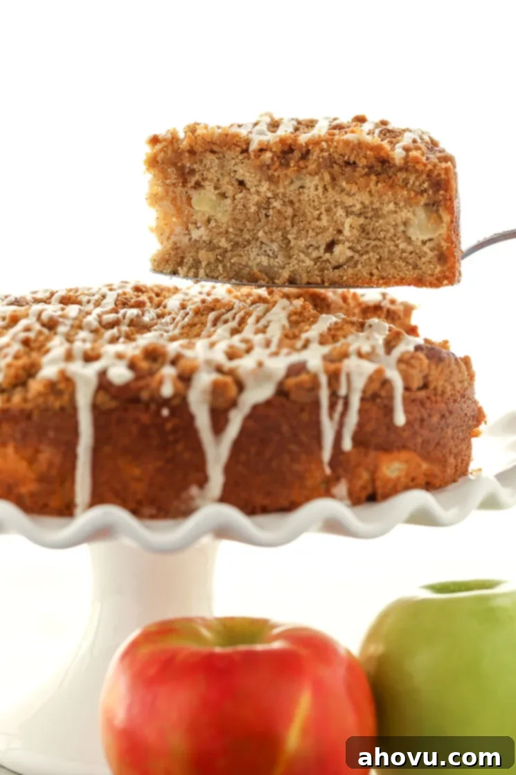 A slice of apple streusel coffee cake being lifted from a cake stand. Two apples rest in the foreground. 