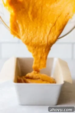 Thick pumpkin bread batter being poured smoothly into a parchment-lined loaf pan, ensuring easy removal after baking.
