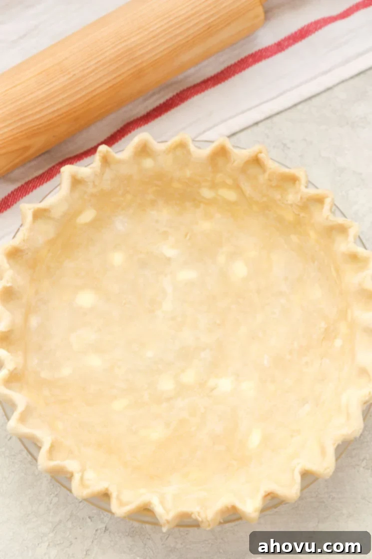 A pie crust in a glass pie dish that's ready to be blind baked. A wood rolling pin and striped towel are next to the crust. 