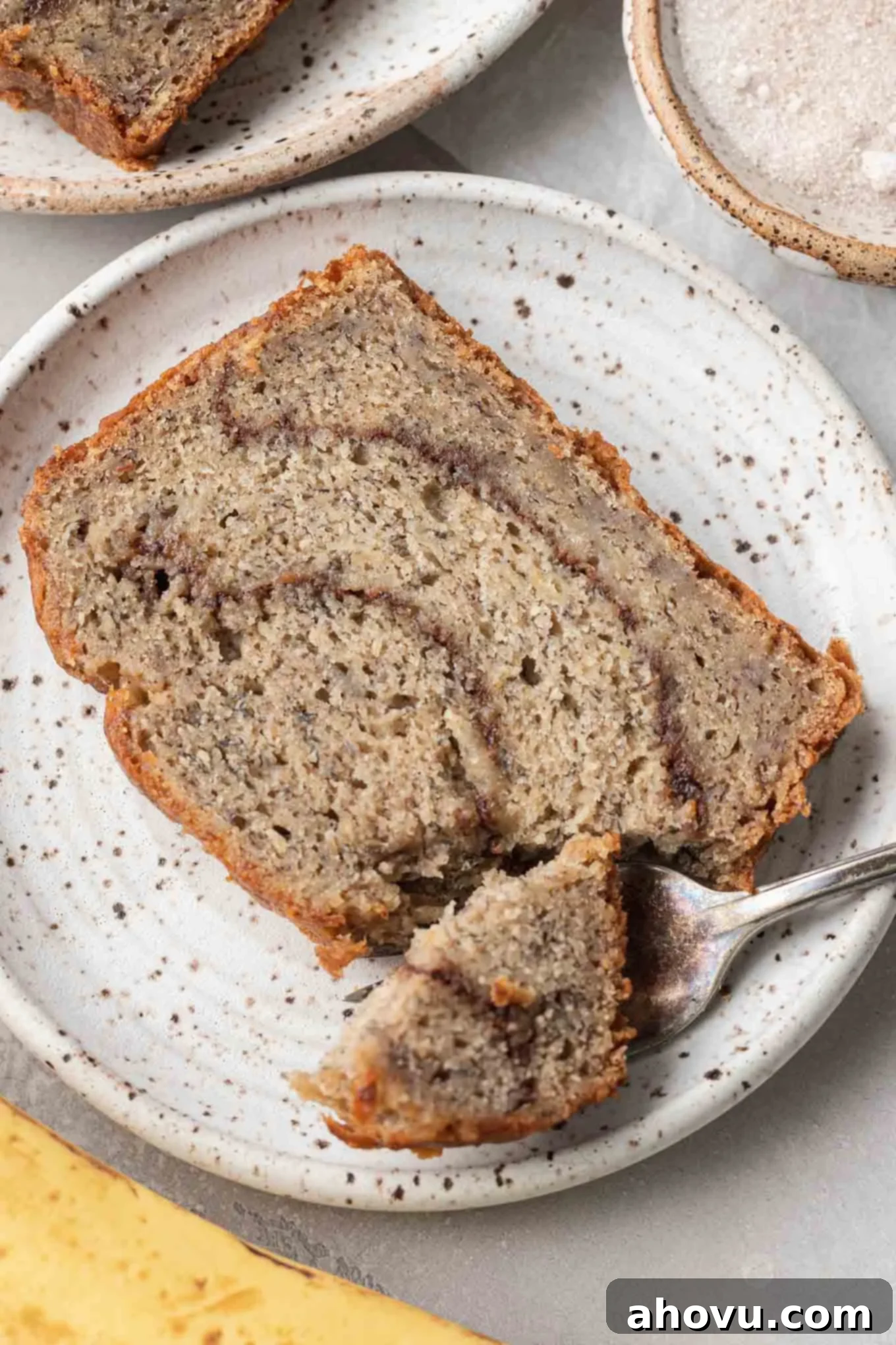 A close up view of a tender slice of cinnamon banana bread on a dessert plate, with a piece speared on a fork, highlighting the moist texture and cinnamon swirl.