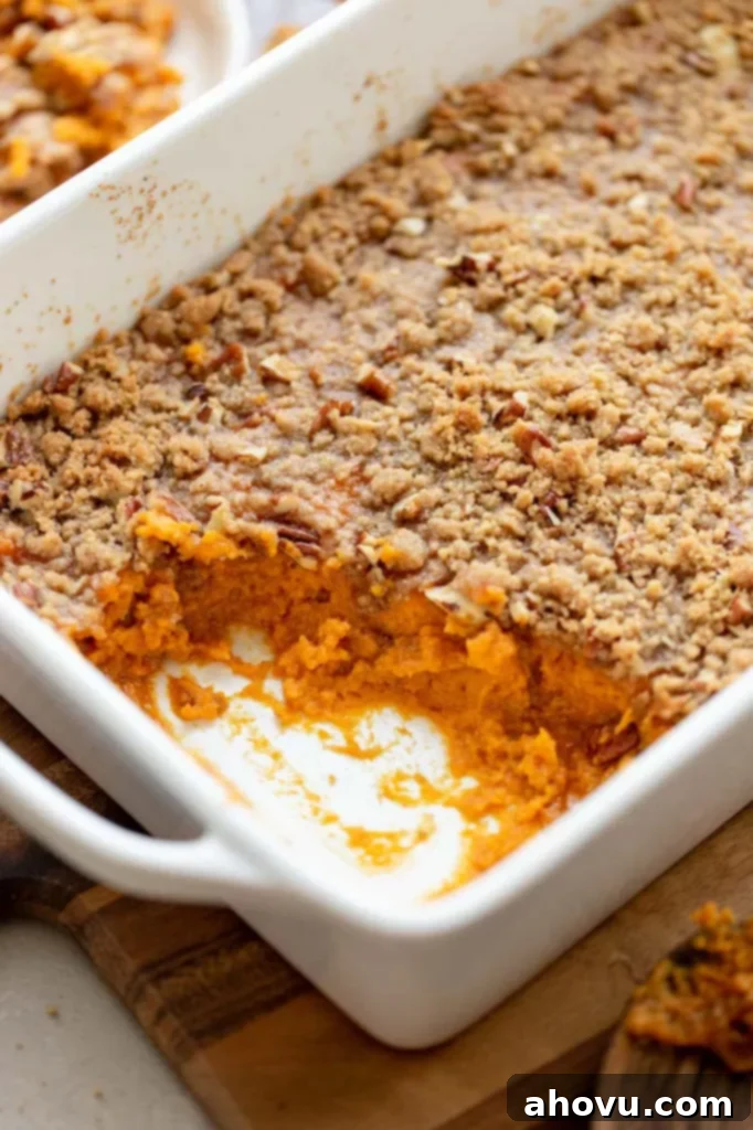 A baked sweet potato casserole in a white baking dish, with a serving removed to show the creamy interior beneath the pecan streusel.