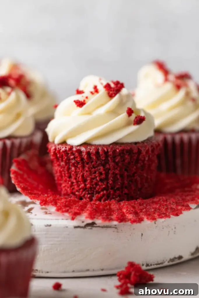 Velvety Red Delights 2 Three beautifully frosted homemade red velvet cupcakes arranged on a pristine white platter. The cupcake in the foreground has its paper liner partially peeled back, revealing its soft, crimson interior and creamy white frosting swirl.