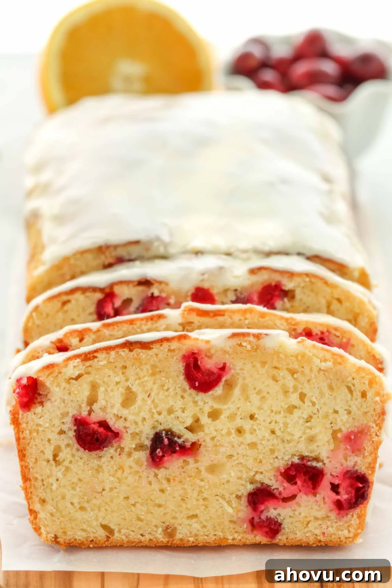 A beautifully baked loaf of cranberry orange bread, richly glazed and garnished with fresh cranberries and orange slices. In the background, half an orange and a bowl of fresh cranberries are visible.