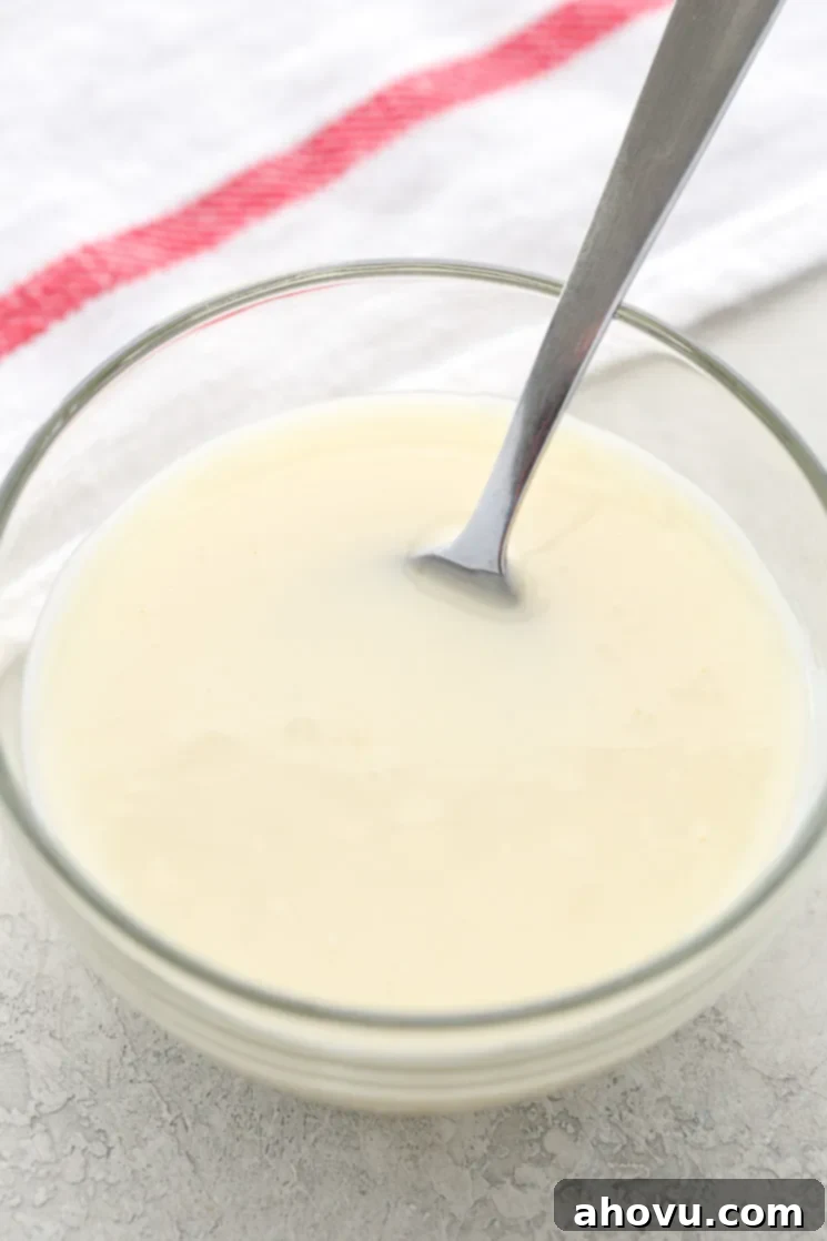 A small, elegant glass bowl filled with a smooth, white vanilla glaze. A silver spoon is gently resting within the bowl, suggesting the glaze is ready to be drizzled over a delicious treat.