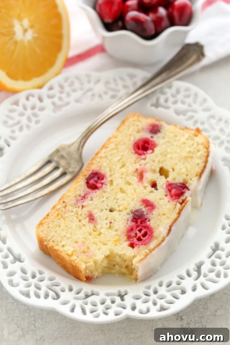 A delightful, perfectly glazed slice of cranberry orange bread rests on a pristine white plate, accompanied by a silver fork. In the soft-focused background, a bowl brimming with fresh, red cranberries and a vibrantly cut half-orange are artfully arranged, subtly hinting at the key ingredients and fresh flavors.