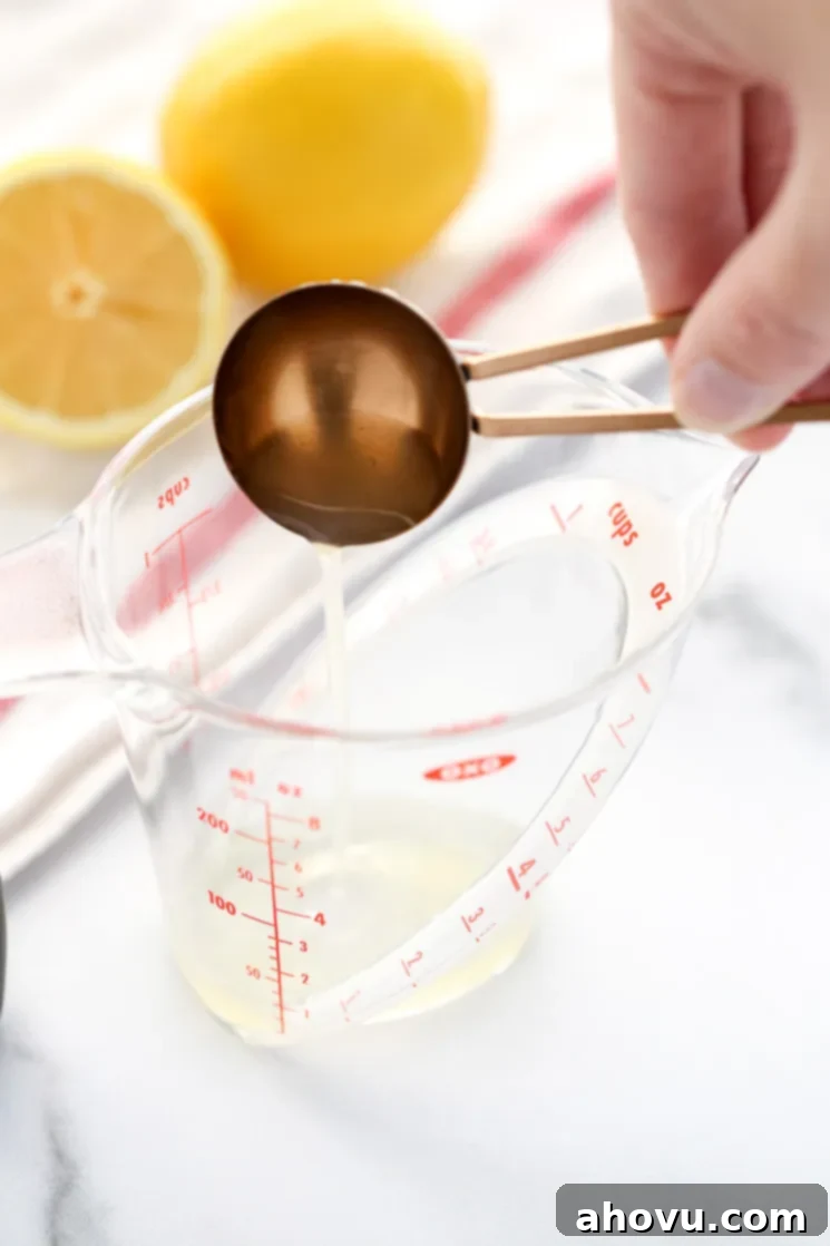 A clear measuring cup with one tablespoon fresh lemon juice, emphasizing the precise measurement of the acid. A white napkin and lemons in the background provide a clean, fresh context.