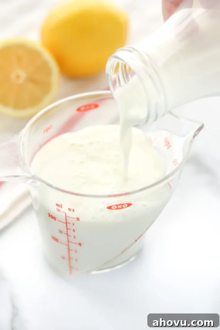 A clear measuring cup being filled with milk, showing the process of combining ingredients. A white napkin and lemons in the background complete the visual scene.