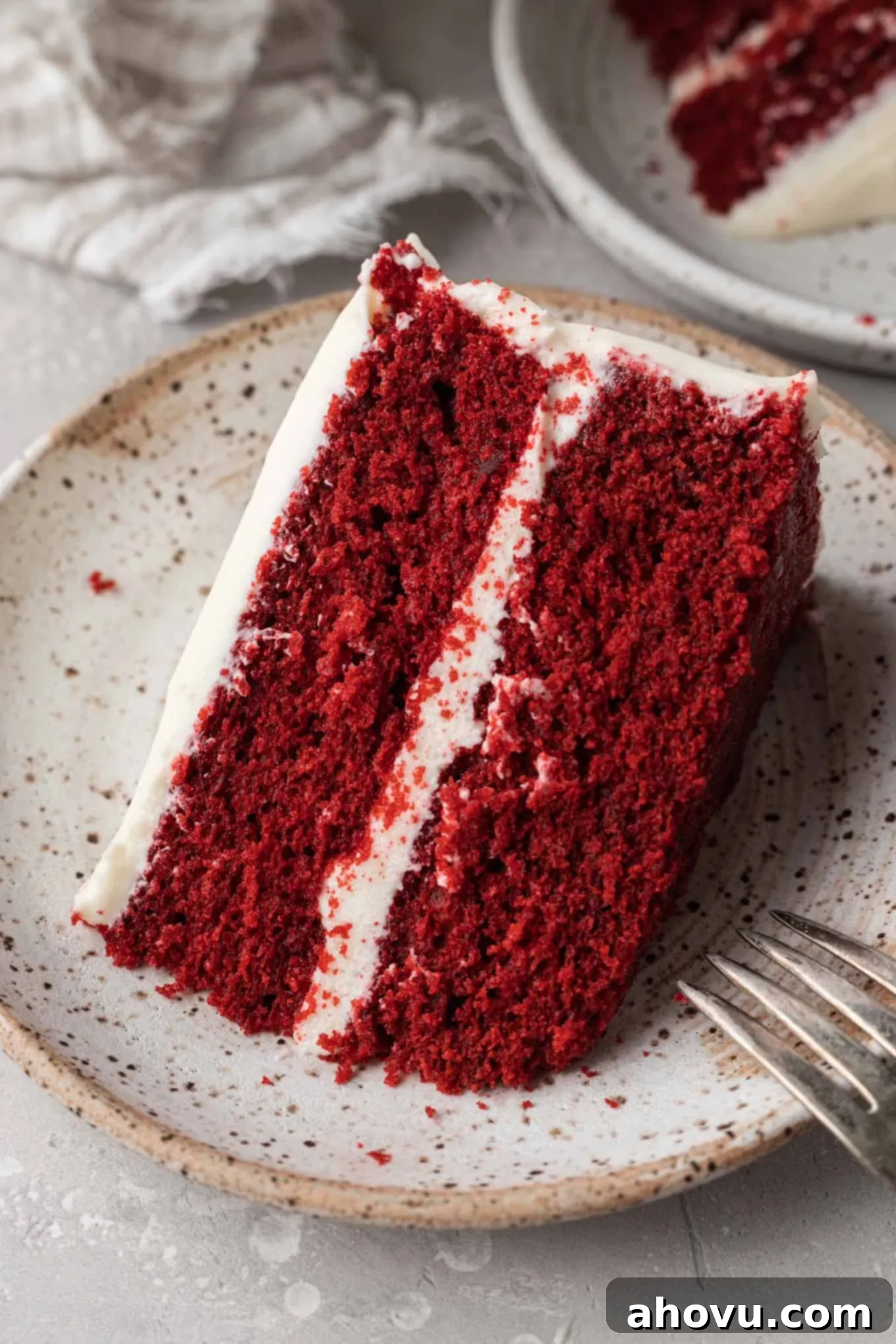A slice of frosted red velvet cake on a speckled dessert plate. 