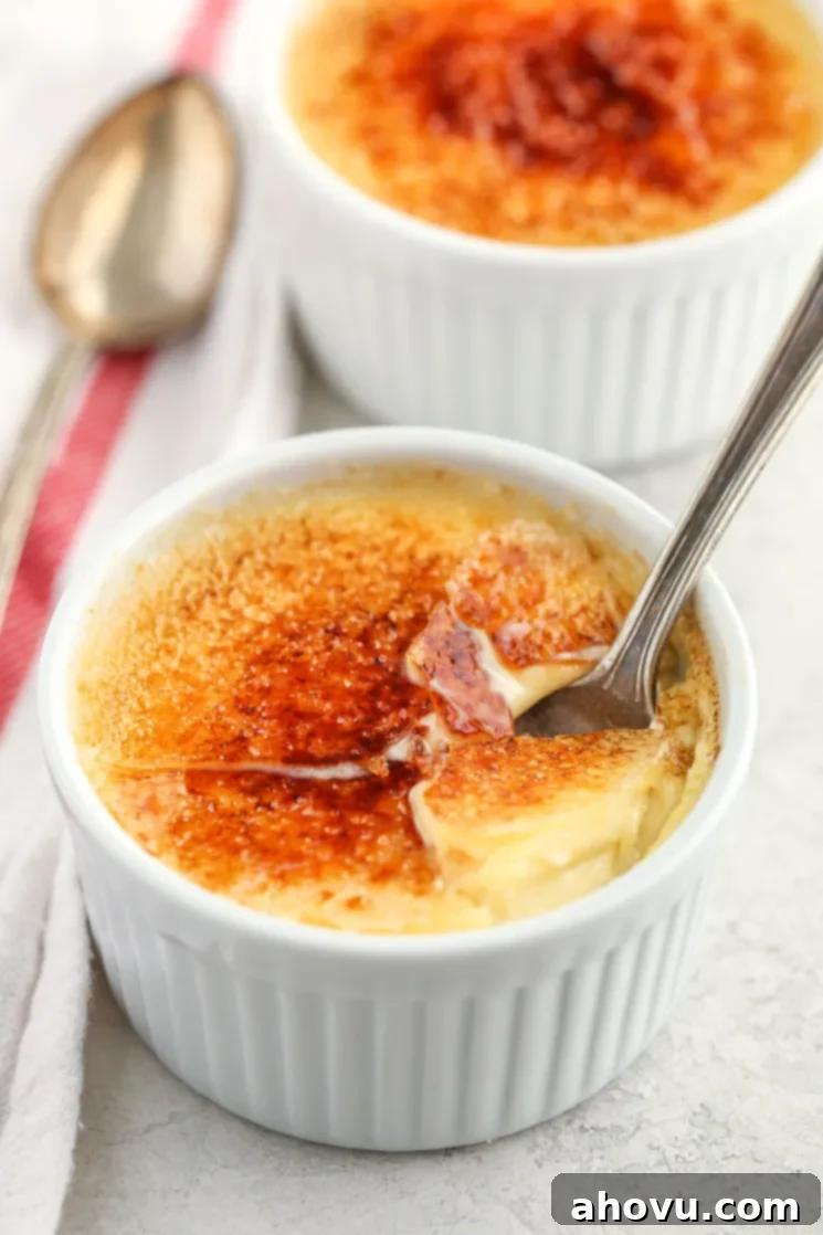 A close-up of a white ramekin filled with crème brûlée and a spoon inside of it along with another crème brûlée and white napkin.