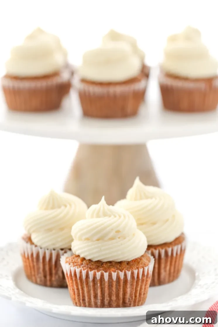 Perfectly Spiced Carrot Delights 5 A decorative white plate holding three carrot cake cupcakes topped with cream cheese frosting in front of a marble cake stand.