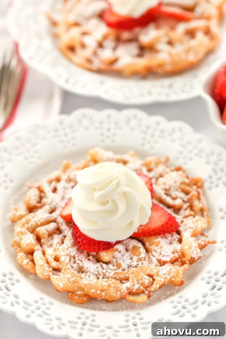 Two funnel cakes topped with powered sugar, strawberries, and homemake whipped cream sitting on decorative white plates. 