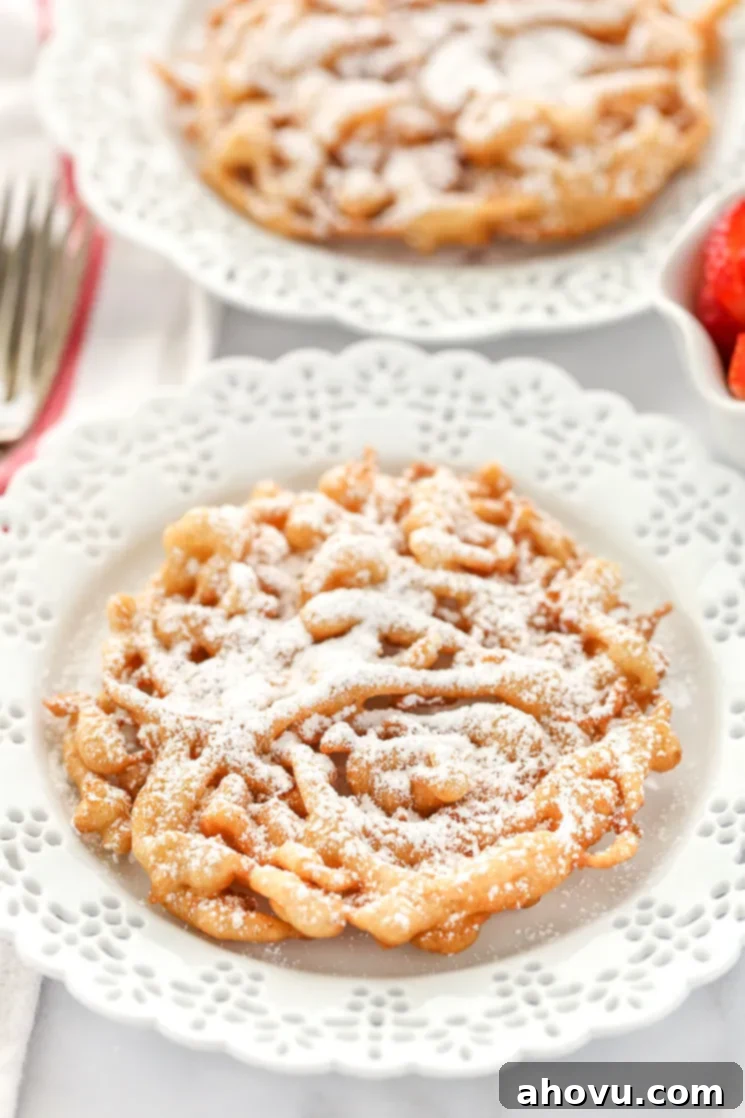 Two homemade funnel cakes topped with powdered sugar on decorative white plates and strawberries on the side. 