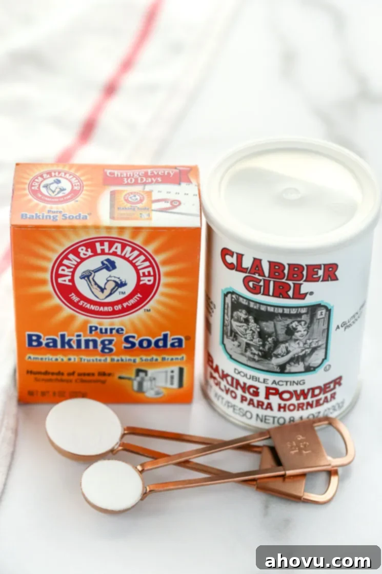 A box of baking soda, can of baking powder, and measuring spoons sitting on top of a marble surface with a white napkin on the side.