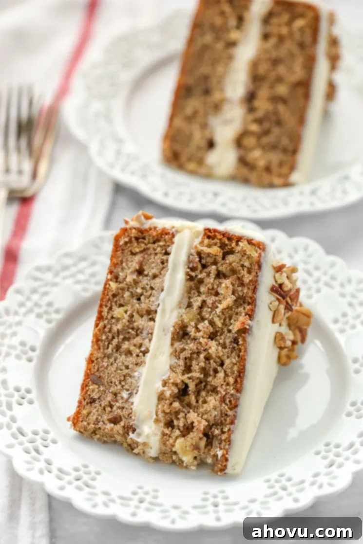Sweet Nectar Cake 2 A beautiful slice of hummingbird cake on a decorative white plate with another slice and a fork subtly blurred in the background, highlighting its moist texture and creamy frosting.