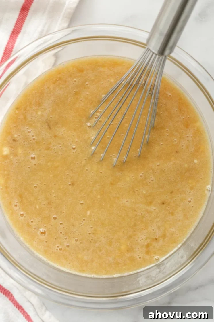 Sweet Nectar Cake 5 A glass mixing bowl brimming with the rich, wet ingredients for a hummingbird cake, featuring mashed bananas, pineapple, and oil, with a metal whisk ready for action.