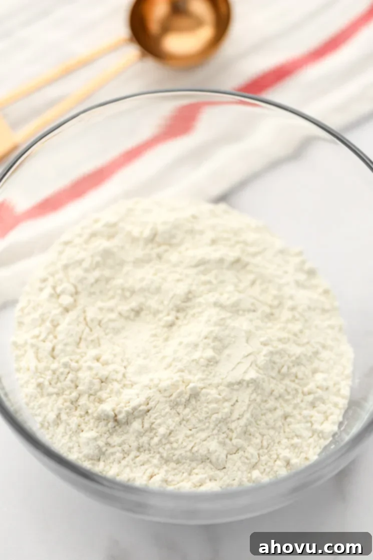 A small glass bowl filled with all-purpose flour and a copper measuring tablespoon in the background.