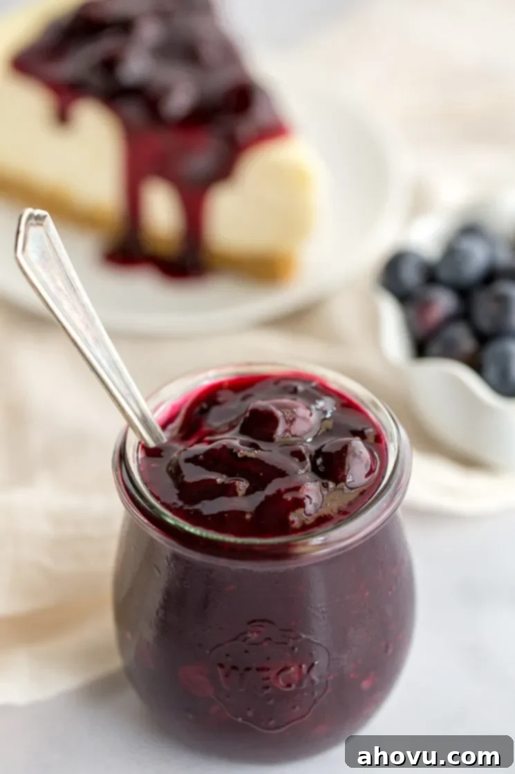 A jar of homemade blueberry sauce with more blueberries and a slice of cheesecake in the background.
