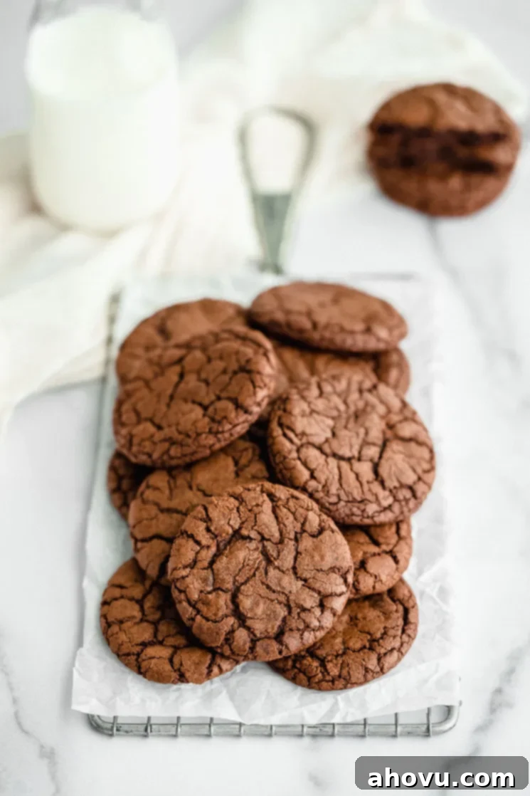 A stack of perfectly baked fudgy brownie cookies on top of an antique safety grater, with more cookies and a glass of milk blurred in the background, highlighting their delicious, crinkly tops.