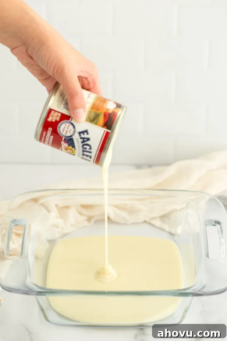 A stream of thick, creamy sweetened condensed milk being poured from a can into a clear glass baking dish.