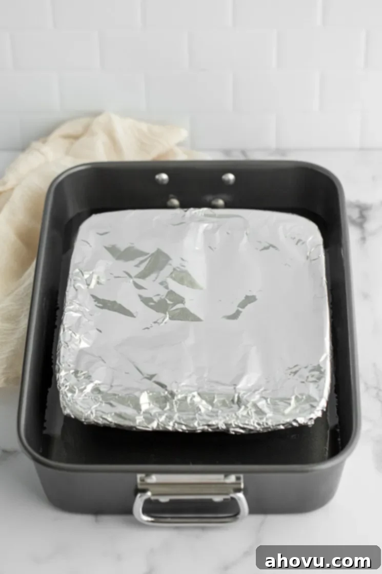 A baking dish filled with sweetened condensed milk, covered tightly with aluminum foil, submerged halfway in hot water within a larger roasting pan, ready for the oven.