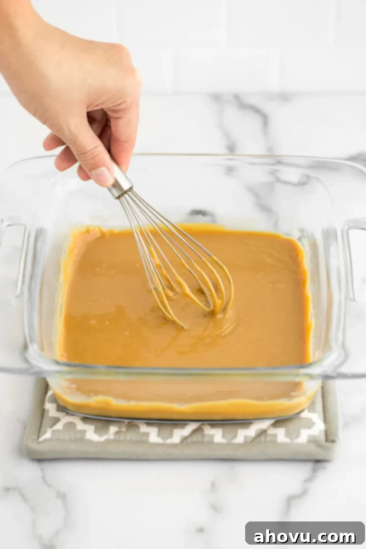 A hand whisking the freshly baked, golden-brown dulce de leche in a clear glass baking dish to achieve a smooth and creamy consistency.