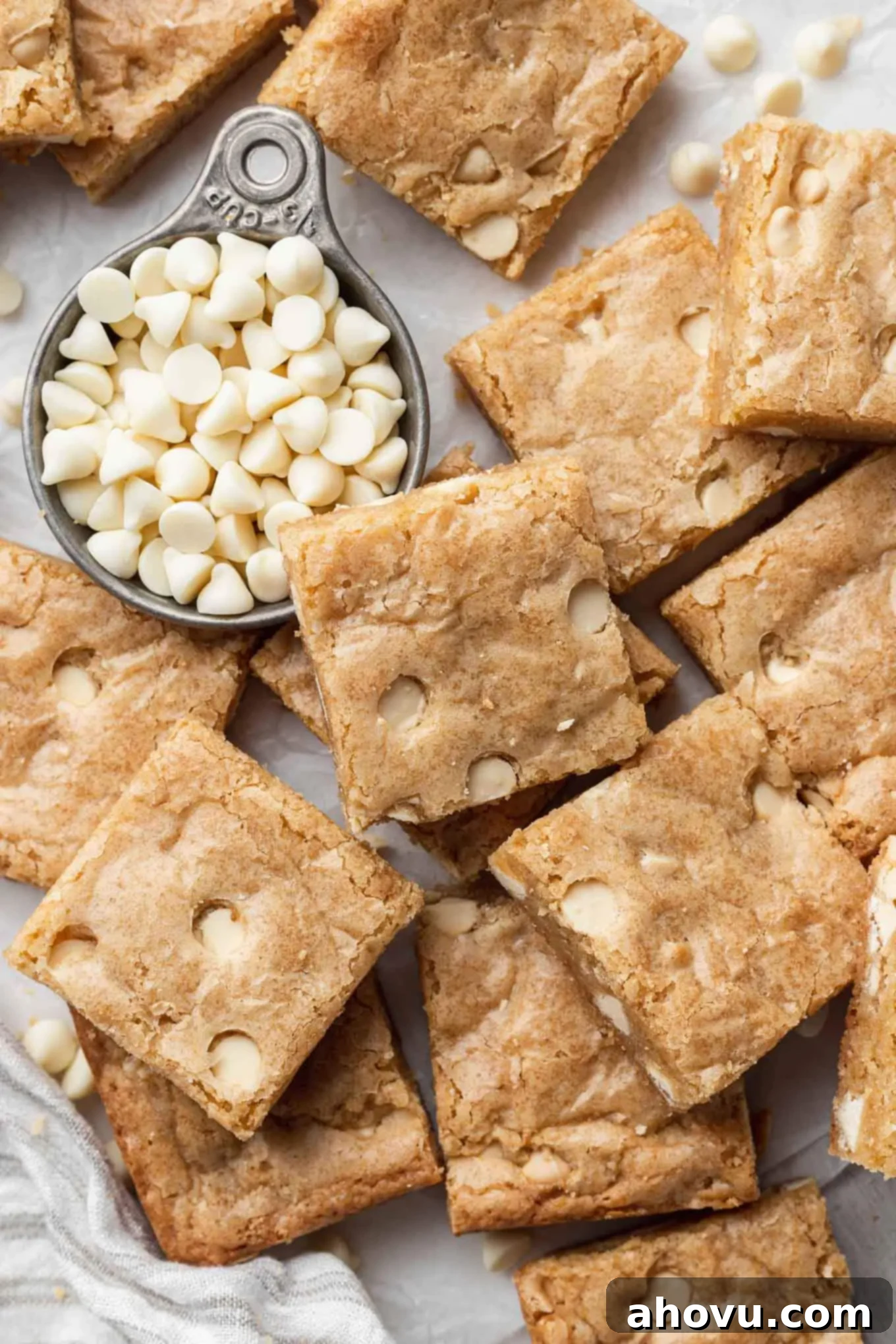 An overhead view of perfectly sliced blondies, some stacked, next to a measuring cup filled with extra white chocolate chips.