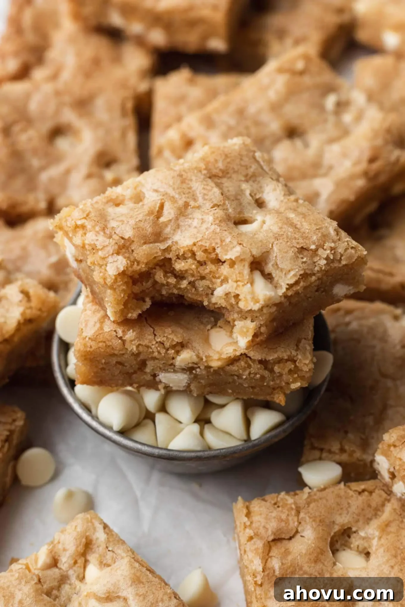 Two golden blondies, one with a bite taken out, neatly stacked atop a small dish of white chocolate chips, highlighting their deliciousness.