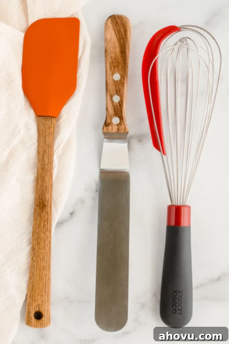 A rubber spatula, an icing spatula, and a whisk resting on top of a marble counter, representing essential baking utensils.