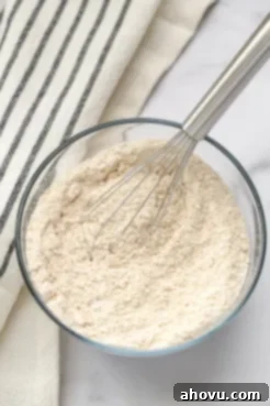 A clear bowl filled with white whole wheat flour, baking powder, baking soda, and salt with a silver whisk.