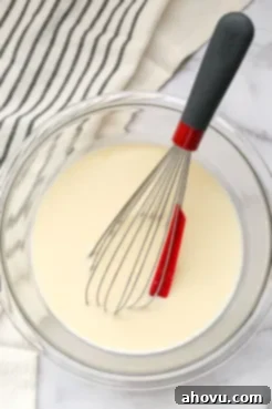 A clear bowl filled with the wet ingredients for pancake batter and a red and black whisk.