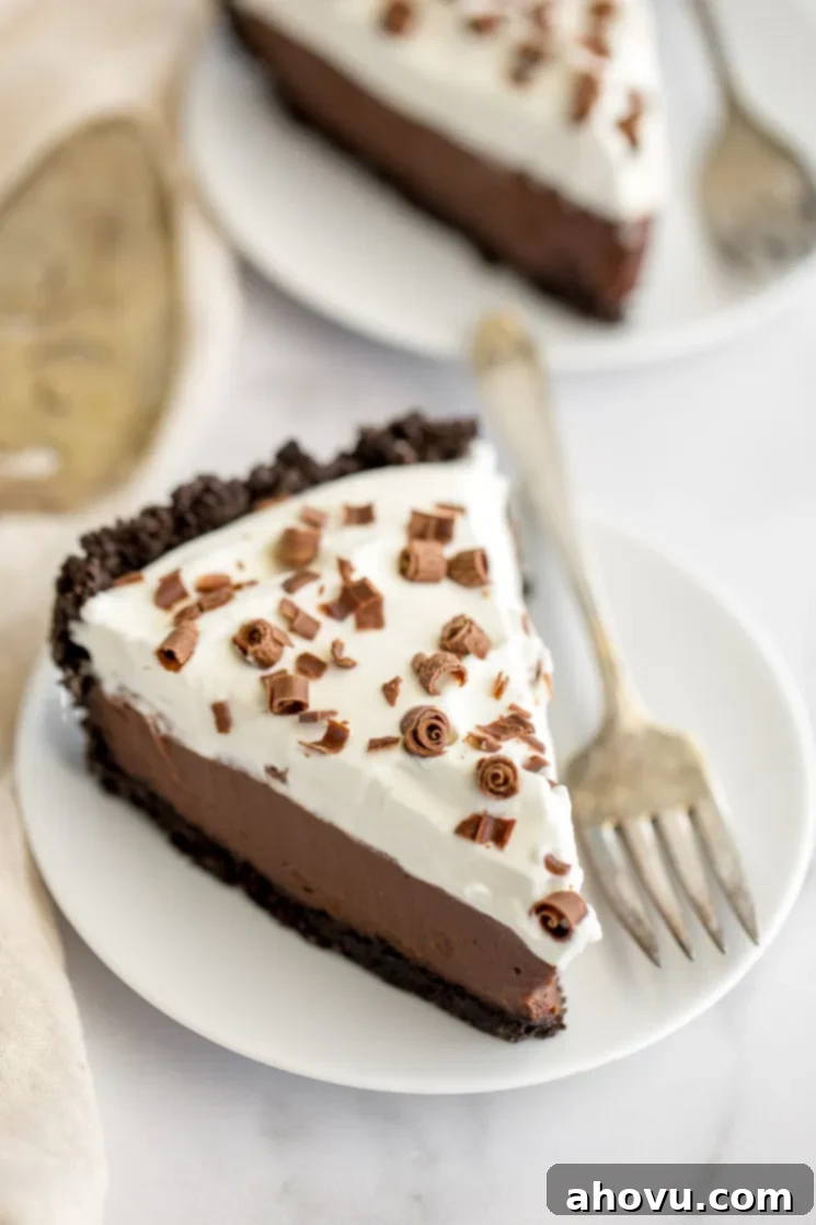 A perfectly sliced piece of homemade chocolate cream pie on a white plate, with the rest of the pie blurred in the background, showcasing its creamy layers.